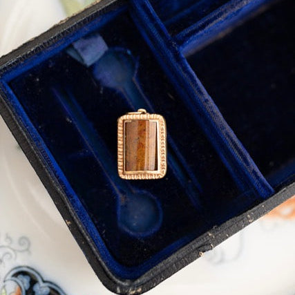 A rectangular gold pendant with a brown striped stone, displayed inside an open dark blue velvet jewelry box on a floral plate.